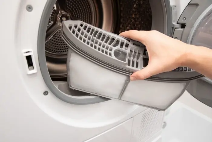 A person removing and cleaning a plastic lint trap filter from a modern front-loading clothes dryer, demonstrating routine maintenance to answer how long should a dryer last in a home and prevent the signs it's time for repair vs. replacement.