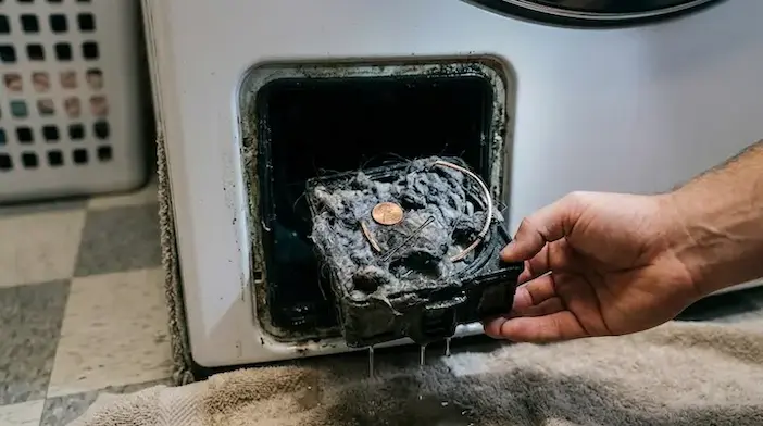 A close-up of a clogged washing machine drain pump filter being pulled out of a white front-load washer in a Salem, Oregon laundry room, covered in lint, hair, and small objects like coins.